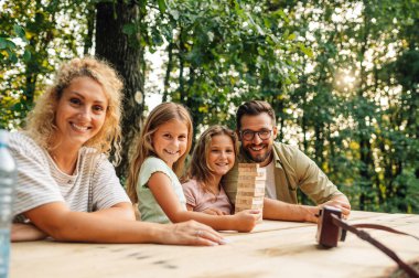 Aile piknik masasında oturuyor ve doğada jenga masası oyunu oynuyor. Neşeli bir aile portresi doğada eğlenceli bir tahta blok oyunu oynarken kameraya gülümsüyor..