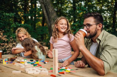 Aile, eğitici oyuncaklarla kaliteli zaman geçirir. Baba gitmek için kahve içiyor ve kız ona gülümsüyor anne ve diğer kız da bulanık arka planda gıdıklanırken. Eğlence zamanı.