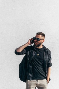 Genç yakışıklı adam akıllı telefon kullanıyor ve dışarıda güneş gözlüğü takıyor. Arka planda beyaz bir duvar var.