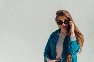 Akıllı telefon kullanan, kot pantolon giyen ve arka planda beyaz bir duvar olan güneş gözlüğü takan güzel bir kadın.