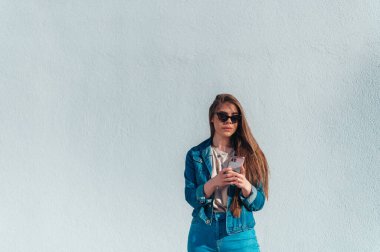 Genç ve güzel bir kadın beyaz bir duvarın önünde dururken akıllı telefon kullanıyor ve güneş tarafından aydınlatılıyor.