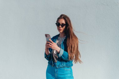 Genç ve güzel bir kadın akıllı telefon kullanıyor ve beyaz bir duvarın önünde dururken kot pantolon ve güneş gözlüğü takıyor.