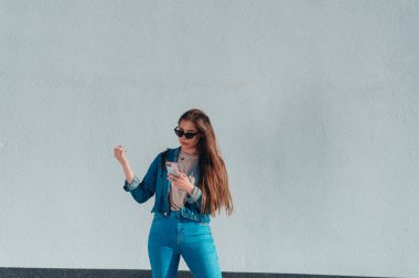 Genç ve güzel bir kadın akıllı telefon kullanıyor ve beyaz bir duvarın önünde dururken kot pantolon ve güneş gözlüğü takıyor.
