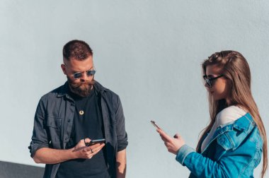Genç, modaya uygun bir çift, beyaz bir duvarın önünde güneş gözlüğü takarken akıllı telefon kullanıyor.