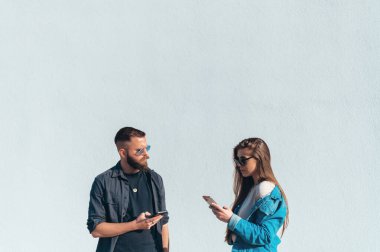 Genç ve güzel bir çift dışarıda beyaz bir duvarın önünde dikilirken akıllı telefon kullanıyor.