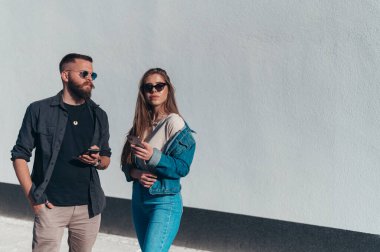 Genç ve güzel bir çift, beyaz bir duvarın önünde dikilip güneşin altında beklerken akıllı telefonlar kullanıyorlar.