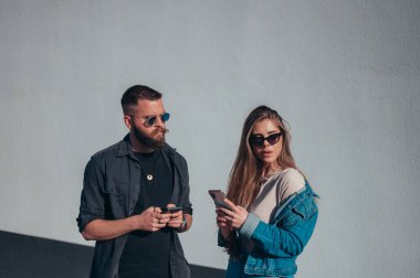 Genç, modaya uygun bir çift, beyaz bir duvarın önünde güneş gözlüğü takarken akıllı telefon kullanıyor.