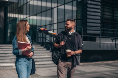 İki genç öğrenci konferansa giderken ellerinde kitap ve kahve paketleri ile fakültenin önünde buluşuyorlar.