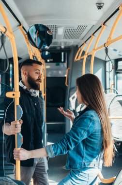 Genç çift hareket halindeki bir otobüste durup sırıkları tutarken konuşuyorlar.