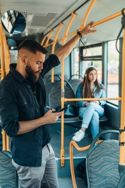 Genç ve yakışıklı bir adam hareket halindeki bir otobüste dikiliyor ve elinde bir çubuk tutuyor.