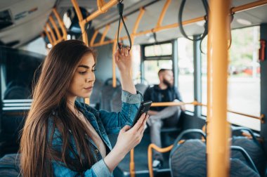 Genç ve güzel bir kadın hareket halindeki bir otobüste sırık tutarken Samrtphone kullanıyor.