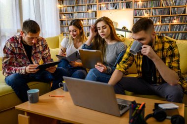 Bir grup genç öğrenci evde birlikte sınava çalışırken bilgisayar ve dijital tablet kullanıyorlar ve kahve içiyorlar.