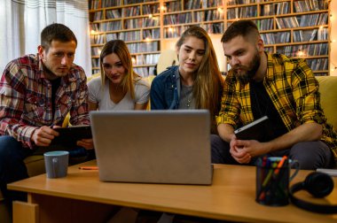 Bir grup öğrenci evde kanepede otururken dizüstü bilgisayar kullanıyor ve sınavlarına birlikte çalışıyorlar.