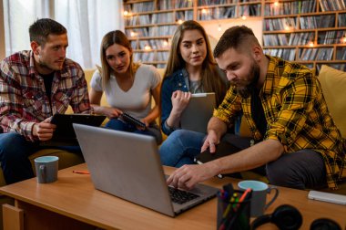 Bir grup öğrenci evde kanepede otururken dizüstü bilgisayar kullanıyor ve sınavlarına birlikte çalışıyorlar.