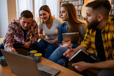 Bir grup genç öğrenci evde birlikte sınava çalışırken bilgisayar ve dijital tablet kullanıyorlar ve kahve içiyorlar.