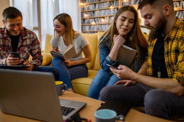 Bir grup genç öğrenci evde birlikte sınava çalışırken bilgisayar ve dijital tablet kullanıyorlar ve kahve içiyorlar.