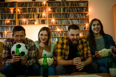 Bir grup arkadaş evde futbol maçı izleyip kanepede dinlenirken bira içerek patlamış mısır yiyor ve televizyon ekranı tarafından aydınlatılıyor.