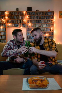 En iyi iki arkadaş evde kanepede dinlenirken video oyunu oynuyorlar ve zafer için bira içip kadeh kaldırıyorlar.