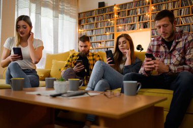 Sıkıcı bir ev partisinde sıcak içecekler içerken akıllı telefonlarına bakan bir grup arkadaş.
