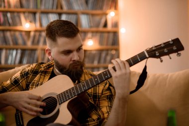 Genç yakışıklı adam evdeki kanepede dinlenirken gitar çalıyor.