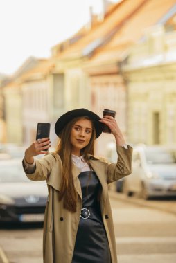 Genç bir kadın akıllı telefonuyla selfie çekiyor ve şehirde yürürken elinde kahve ve şapkasıyla geziyor.