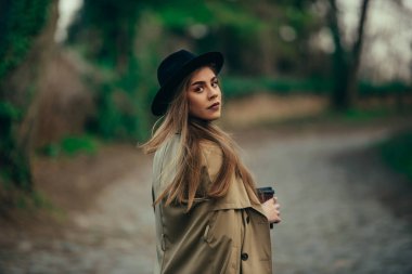 Güzel, modaya uygun bir kadın doğada yürürken ve kahve içerken omzunun üstünden bakıyor.