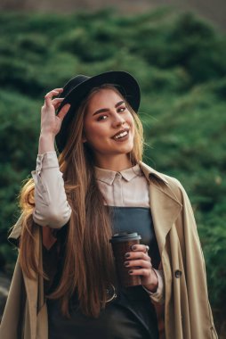 Güzel bir kadın elinde şapkayla gülümsüyor ve kahve ısmarlıyor.