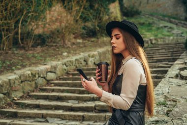 Kahve içerken, deri bir elbise ve şapka giyerken akıllı telefon kullanan genç bir kadının fotoğrafı.