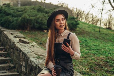 Akıllı telefonu tutarken gülümseyen ve parkta bir duvarda oturan çekici bir genç kadının görüntüsü.