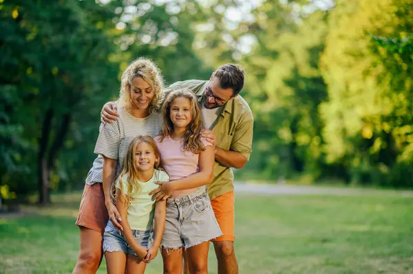 Neşeli bir aile kameraya gülümserken doğada sarılıyor ve ayakta duruyor. Güneşli bir yaz gününde, doğadaki ormanın yakınındaki çayırlarda çocuklarını kucaklayan ebeveynlerin portresi. Aile fotoğrafı..