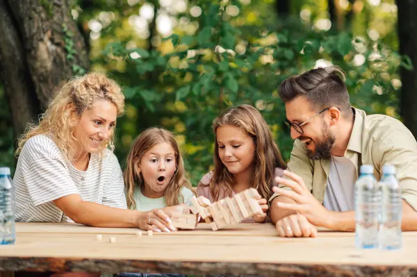 Dört kişilik mutlu aile, doğada masada otururken tahta bloklarla jenga oyunu oynuyor. Aileler ve kızları doğada masada oturup, eğleniyorlar ve tahta blok yapboz oynuyorlar..