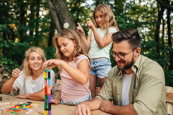 Neşeli bir aile piknik için doğada masada oturuyor ve ahşap fayanslardan sağlam bir yapı inşa ediyor. Eğlenceli bir aile renkli ahşap kiremitlerle oynuyor. Takım çalışması, masa oyunları konsepti.