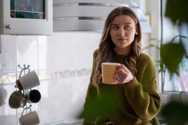 Çekici genç bir kadın mutfakta duruyor ve bir fincan çay içiyor. Rahatlama anı. Mutfakta sabah kahvesinin tadını çıkarıyor..