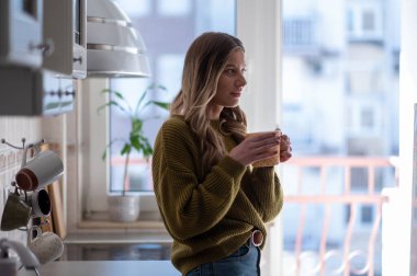 Çekici genç bir kadın evde mutfakta dikilirken bir fincan çay içiyor. Evde boş zaman aktivitesi kavramı.