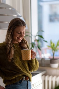 Çekici genç bir kadın mutfakta duruyor ve bir fincan çay içiyor. Rahatlama anı. Mutfakta sabah kahvesinin tadını çıkarıyor..
