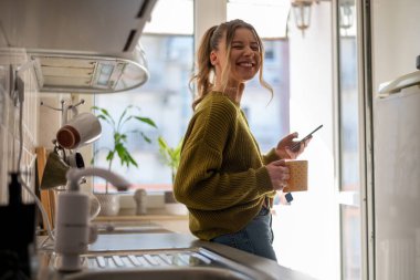 Evdeki mutfakta kahve içerken akıllı telefonunu kullanan gülümseyen genç bir kadın. Sabah konseptinde. Gülümseyen kadın telefon mesajı okuyor..