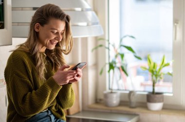 Genç bir kadın, sabahları evdeki parlak bir mutfakta akıllı telefon kullanıyor. Evde keyifli bir sabah..