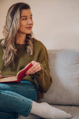 Evdeki kanepede otururken kitap okuyan güzel bir kız. Ev ve konfor kavramları.