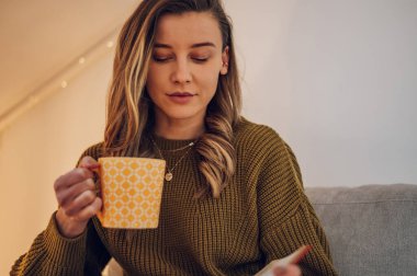En sevdiği romanı okurken ve çay içerken oturma odasındaki kanepede dinlenen güzel genç kadın. Evde kendimi rahat hissediyorum..