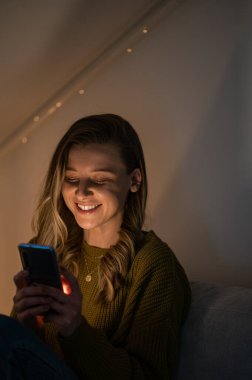 Çekici genç bir kadın evdeki kanepede otururken akıllı telefon kullanıyor ve cihaz ekranı tarafından aydınlatılıyor. İletişim ve rahatlık kavramı.