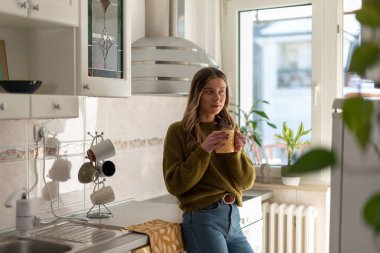 Çekici genç bir kadın evde mutfakta dikilirken bir fincan çay içiyor. Evde boş zaman aktivitesi kavramı.