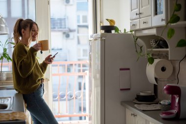 Evdeki mutfakta kahve içerken akıllı telefonunu kullanan gülümseyen genç bir kadın. Sabah konseptinde. Gülümseyen kadın telefon mesajı okuyor..