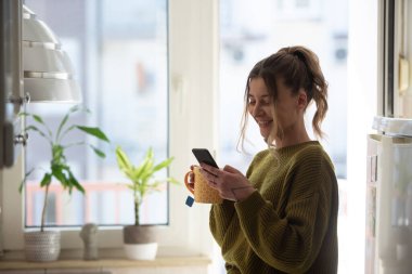 Genç bir kadın sabahları çay içiyor ve evdeki parlak bir mutfakta akıllı telefon kullanıyor. Evde keyifli bir sabah..