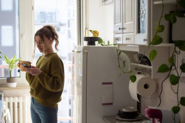 Genç bir kadını çay içerken ve işe gitmeye hazırlanırken mutfağında akıllı telefon kullanırken çekmek. Evde eğlenirken video izlemek ya da arkadaşlarla mesajlaşmak..