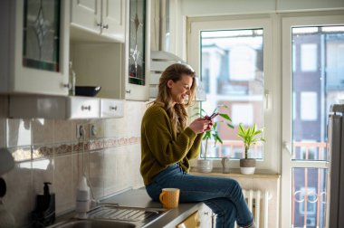 Genç bir kadın sabahları çay içiyor ve evdeki parlak bir mutfakta akıllı telefon kullanıyor. Evde keyifli bir sabah..