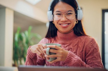 Genç Vietnamlı kadın ev ofisinde çalışıyor ve müzik dinlerken, kahve ya da çay içerken dizüstü bilgisayar kullanıyor. Esnek çalışma alanı ve çalışma zamanı. Güzel Asyalı iş kadını ya da öğrenci.