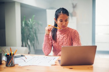 Genç Asyalı iş kadını evde dizüstü bilgisayarda çalışıyor ve projeyi düşünürken elinde kalem tutuyor. Gülümseyen Vietnamlı kadın uzaktan iletişim kurmaktan zevk alıyor. Evden çalışıyorum. Boşluğu kopyala.