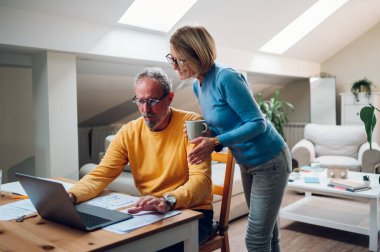 Evde dizüstü bilgisayar kullanan orta yaşlı mutlu bir çiftin portresi. Gülümseyen yaşlı aile haberleri okuyor ya da internetten alışveriş yapıyor. Yaşlılar ve teknoloji kavramı. Boşluğu kopyala.