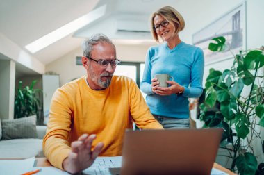 Üst düzey orta yaşlı mutlu çift evde dizüstü bilgisayar kullanıyor. Gülümseyen yaşlı aile haberleri okuyor ya da internetten alışveriş yapıyor. Yaşlılar ve teknoloji kavramı. Bir adama odaklan..