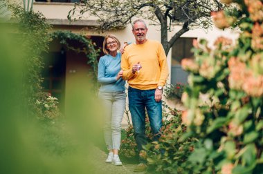 Mutlu, olgun bir çift yeni evlerini satın aldı ve önünde durup anahtarlarını gösterdi. Gayrimenkul sahipleri. Yeni aile evlerinin önünde poz veren yaşlı evli bir çift. Kameraya bakıyorum..
